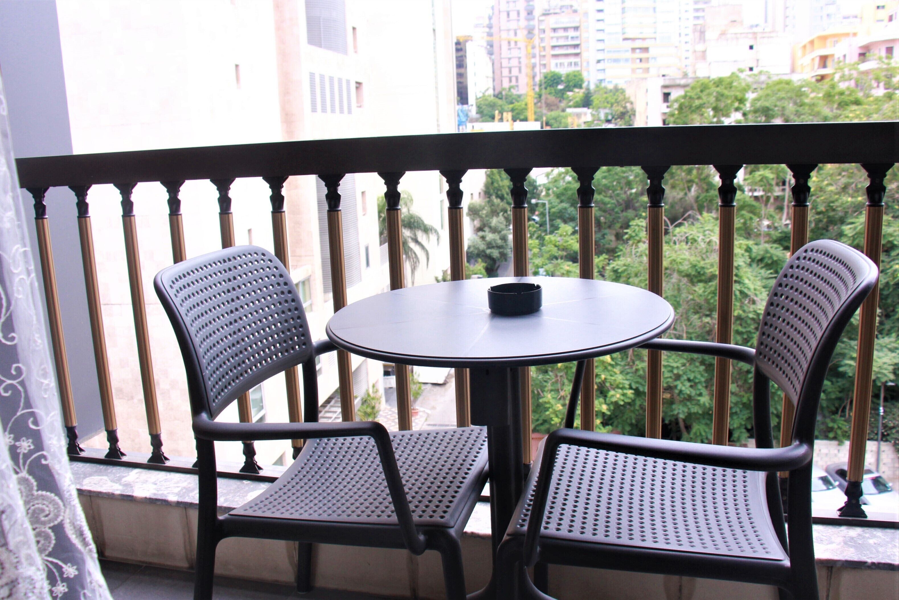 Un balcón con dos sillas y una mesa, con vista a un paisaje urbano con edificios y árboles.