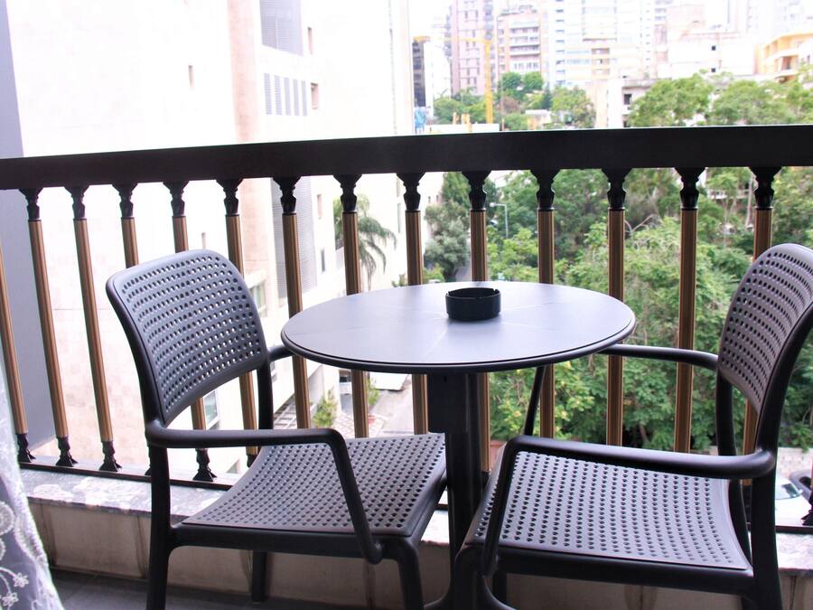 Un balcón con dos sillas y una mesa, con vista a un paisaje urbano con edificios y árboles.