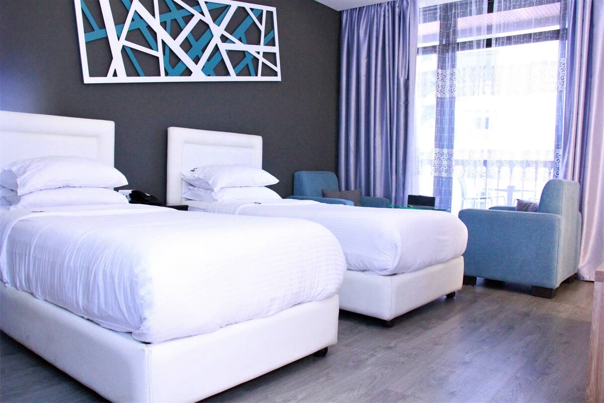A hotel room with two beds, a blue armchair, and a large window with curtains.