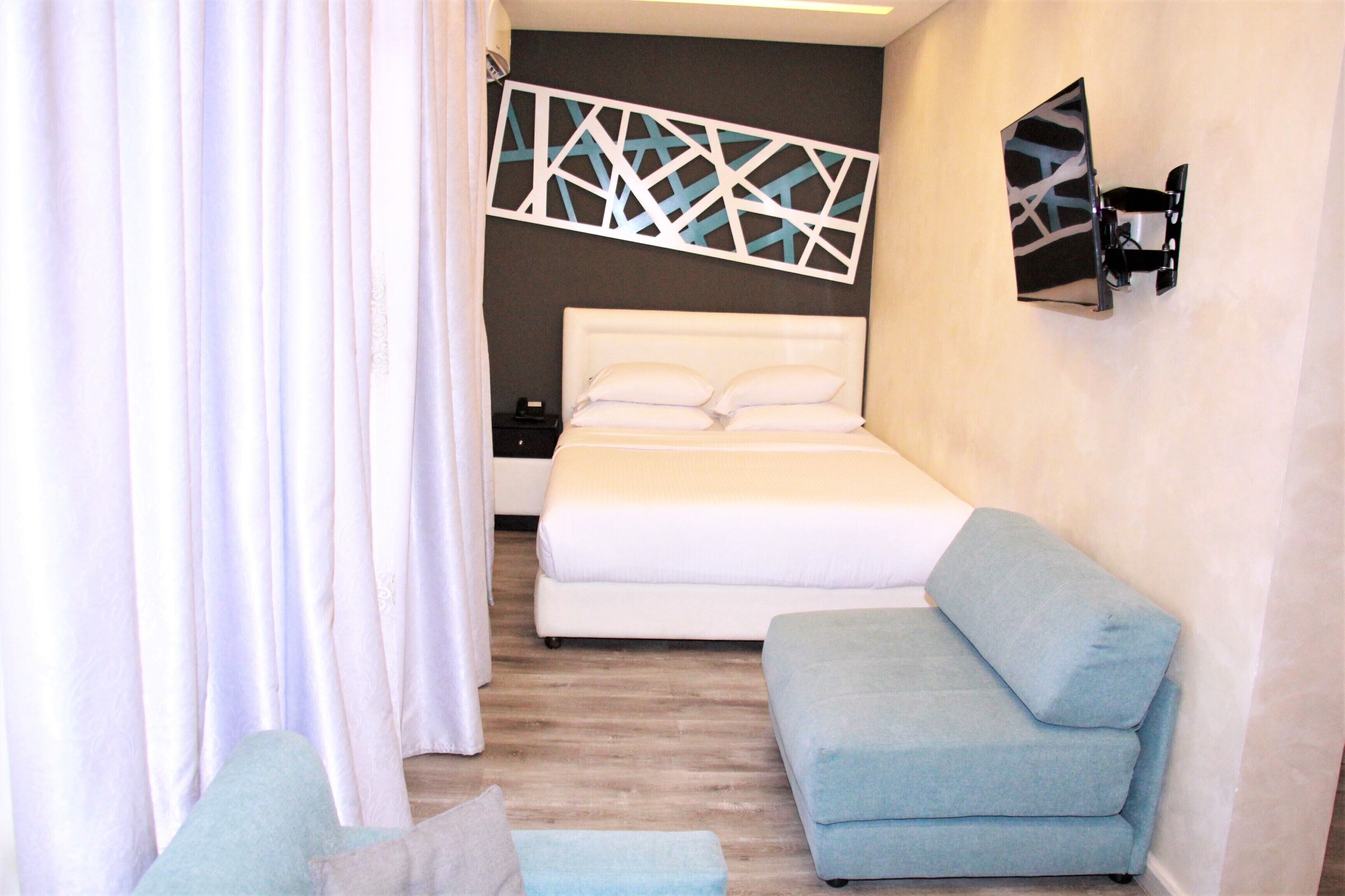 Un dormitorio con cama, sofá azul, mesita de noche y televisión instalada en la pared.