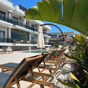 A poolside area with wooden lounge chairs, a white umbrella, and a building with balconies and a 'S' sign. - Suena Hotel (Cesme)