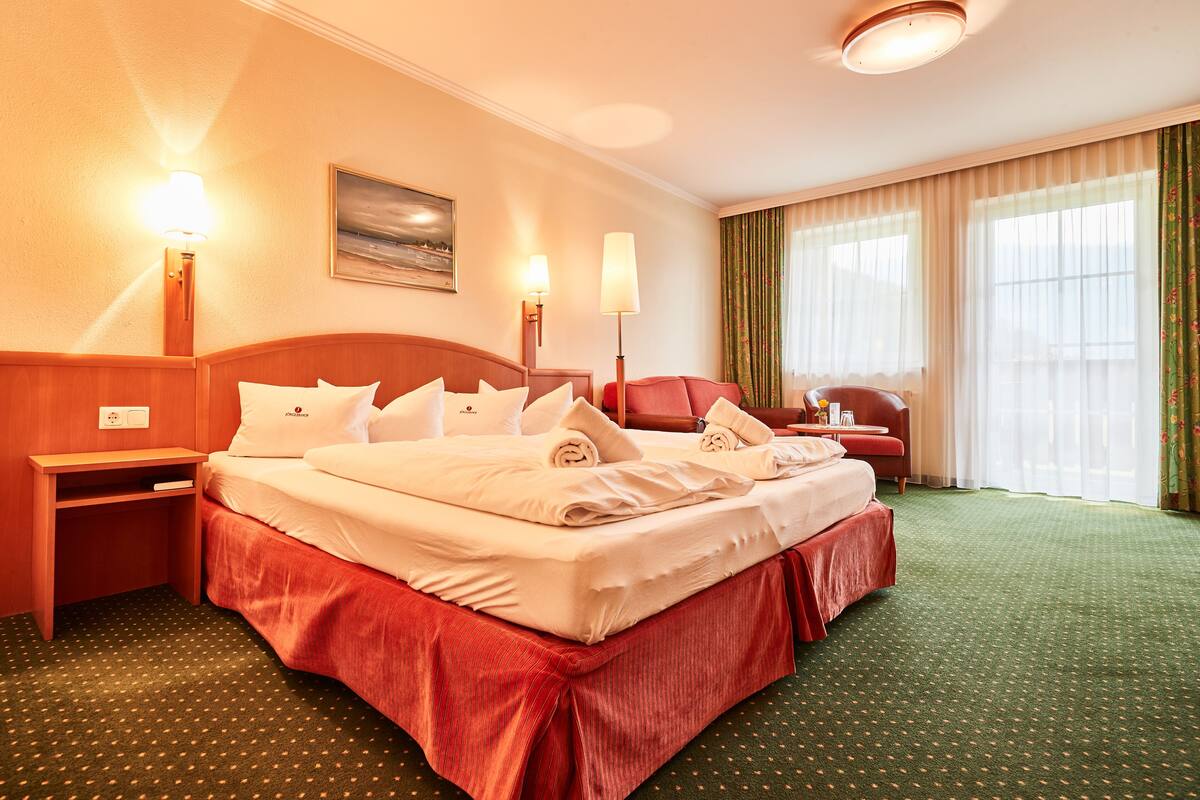 A hotel room with a large bed, two bedside lamps, a red chair, and a view of the outdoors through the windows.