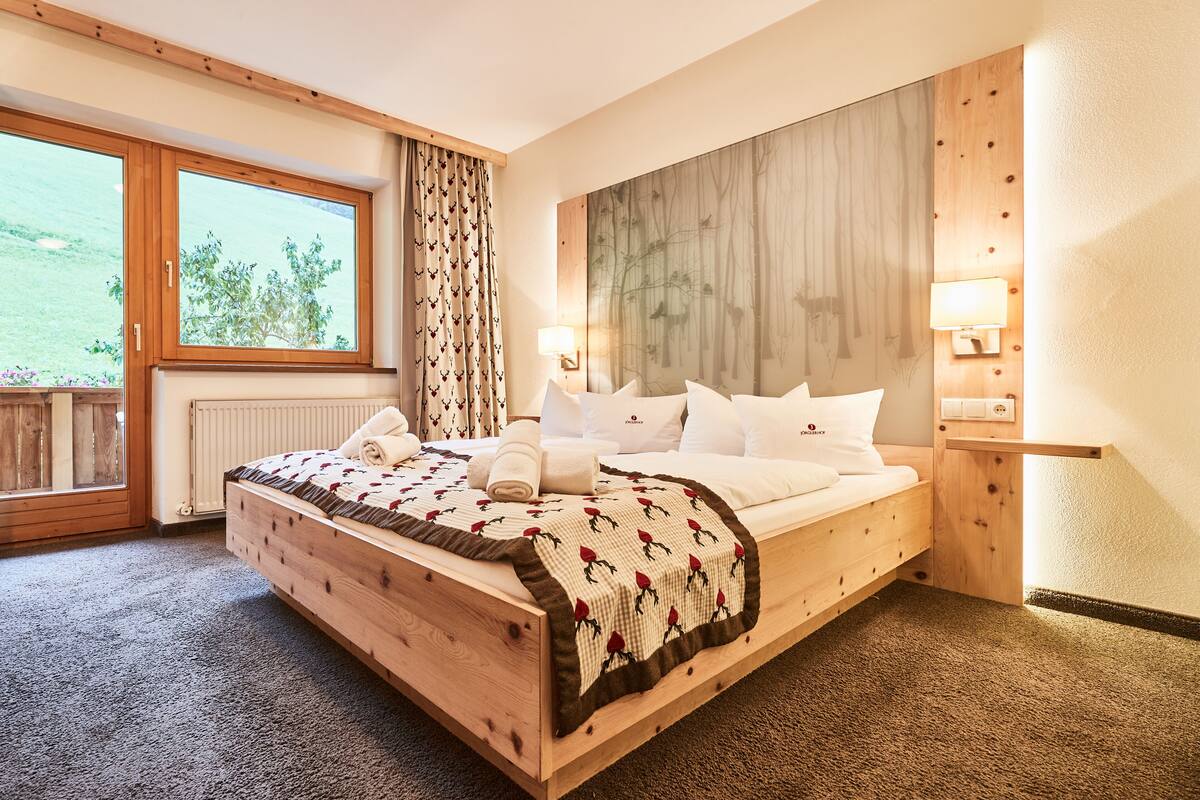 A bedroom with a wooden bed, a large window, a view of greenery, and a wall-mounted lamp.