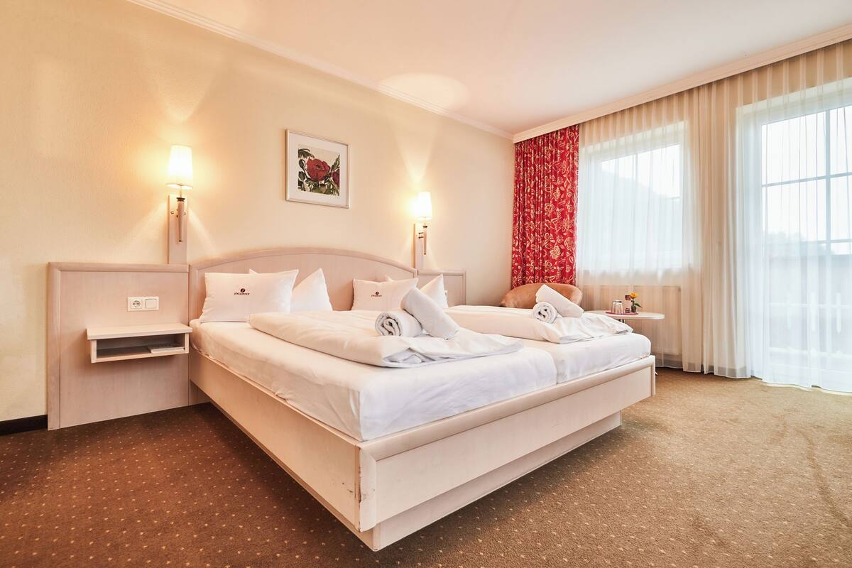 A double bed with white linens, two bedside tables, a nightstand, a wall-mounted lamp, a framed picture, and a window with sheer curtains.