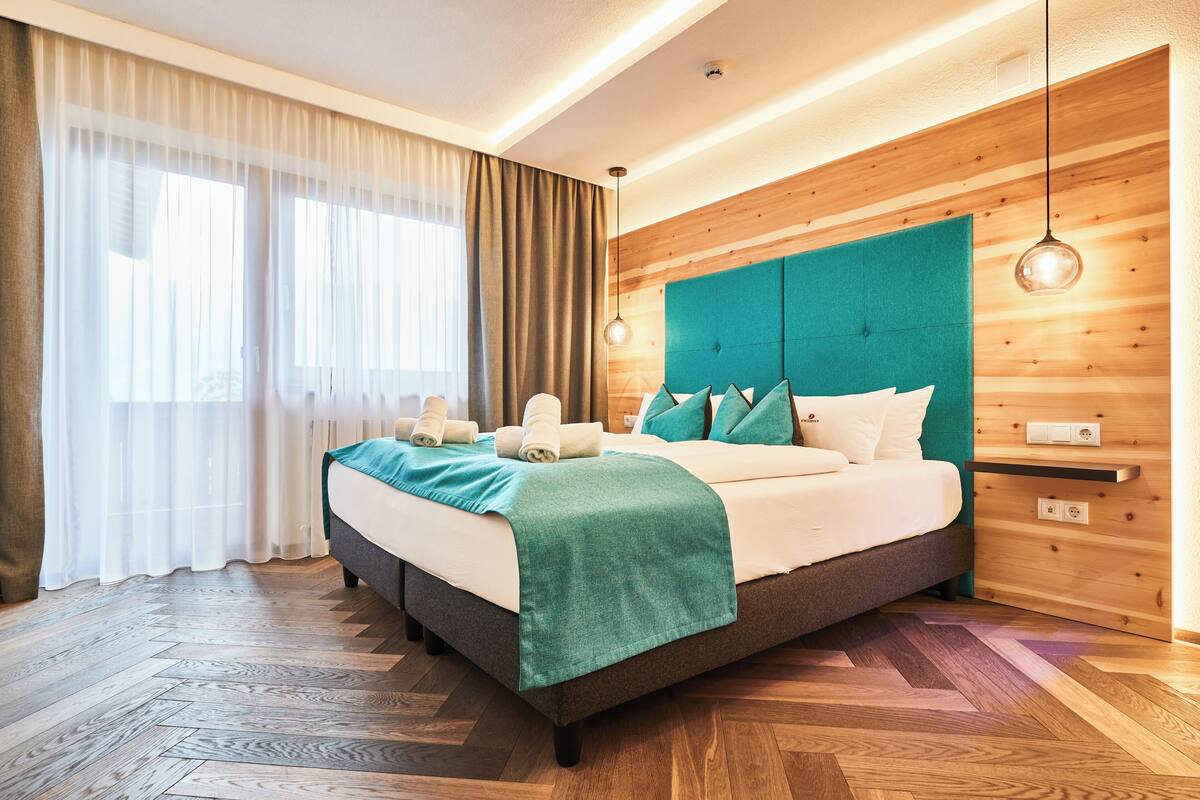A modern hotel room with a large bed, wooden panel walls, a herringbone floor pattern, and a large window with sheer curtains.