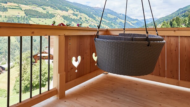 Un balcón de madera con una silla de mimbre colgante, barandillas de madera y vistas a colinas verdes.