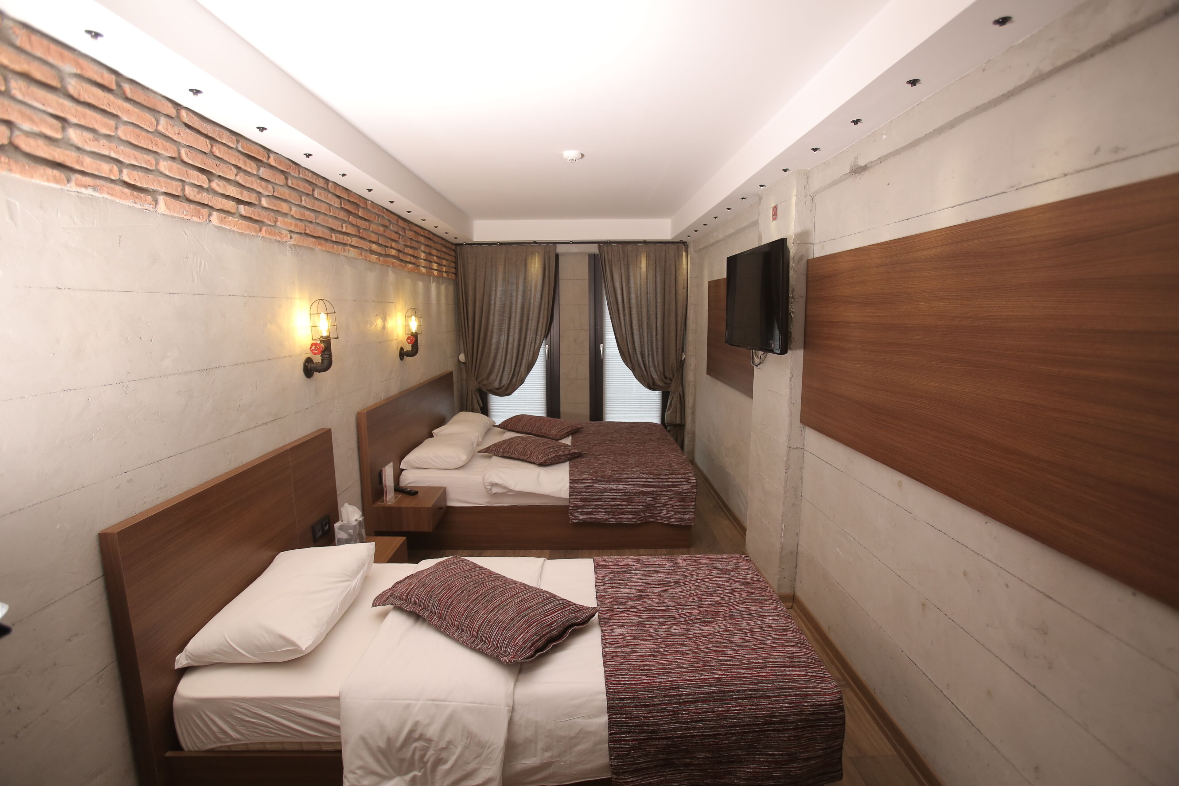 A hotel room with two beds, a brick wall, a flat-screen TV, and a window with curtains.