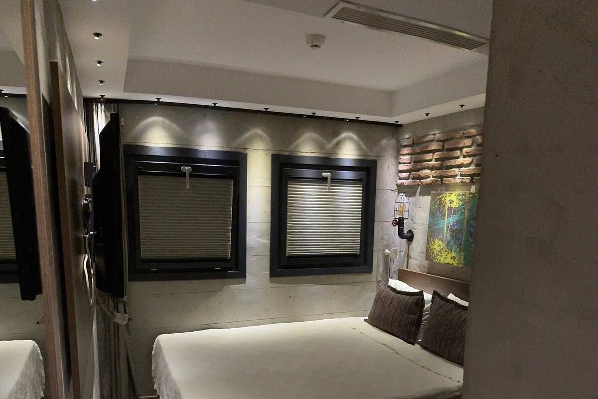 A compact bedroom with a bed, two windows with blinds, a wall-mounted light fixture, and a partial view of a bathroom.