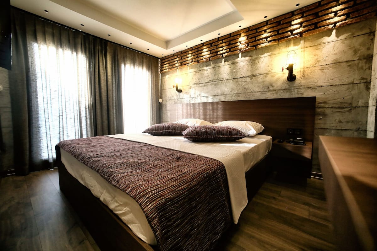 A modern hotel room with a large bed, dark wood flooring, a brick-patterned headboard, and a large window with curtains.