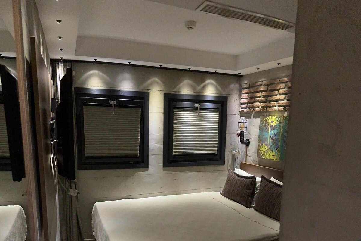 A compact bedroom with a bed, two windows with blinds, a wall-mounted light fixture, and a partial view of a bathroom.