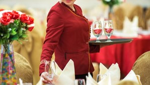 A person in a red uniform serving at a table with red tablecloths, white napkins, and a vase of red flowers.
