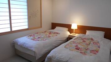 A room with two single beds, each with a floral bedspread and white bedding. There is a nightstand with a lamp between the beds, and a window with blinds on the left.