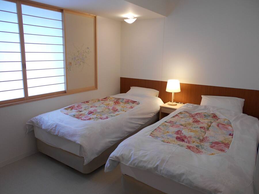 A room with two single beds, each with a floral bedspread and white bedding. There is a nightstand with a lamp between the beds, and a window with blinds on the left.