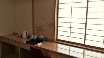 A hotel room with a desk, a red chair, a large window with a grid pattern, and a wall with floral artwork.