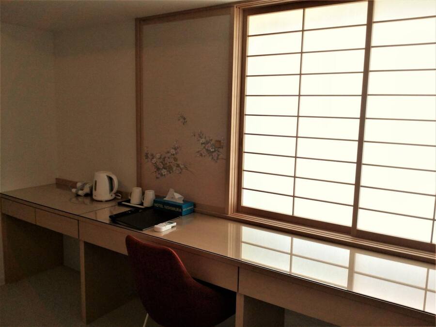 A hotel room with a desk, a red chair, a large window with a grid pattern, and a wall with floral artwork.