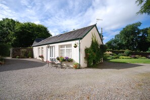 A white building with a gravel driveway, a small table and chair outside, and a garden with various plants and flowers. - Bonawe House Holiday Cottages (Taynuilt)
