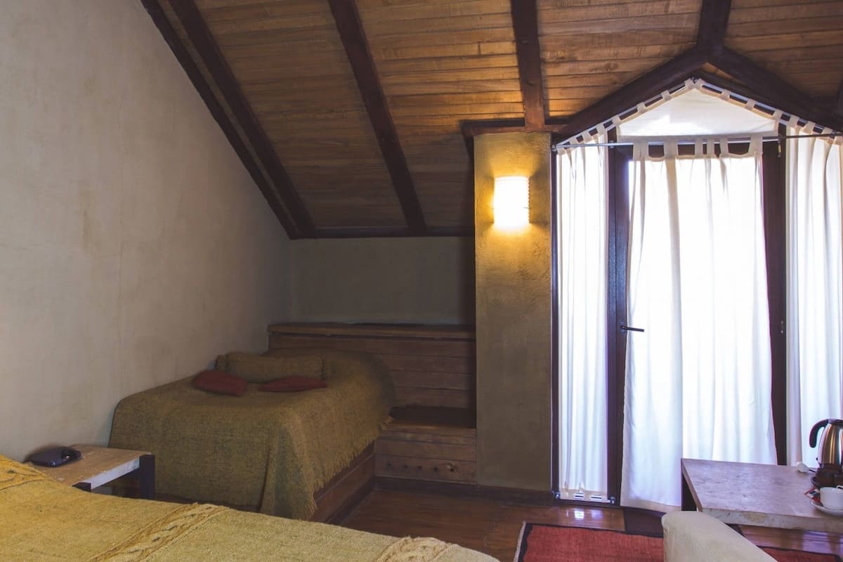 A rustic room with wooden beams, a bed with a red pillow, a table with a kettle, and a window with curtains.
