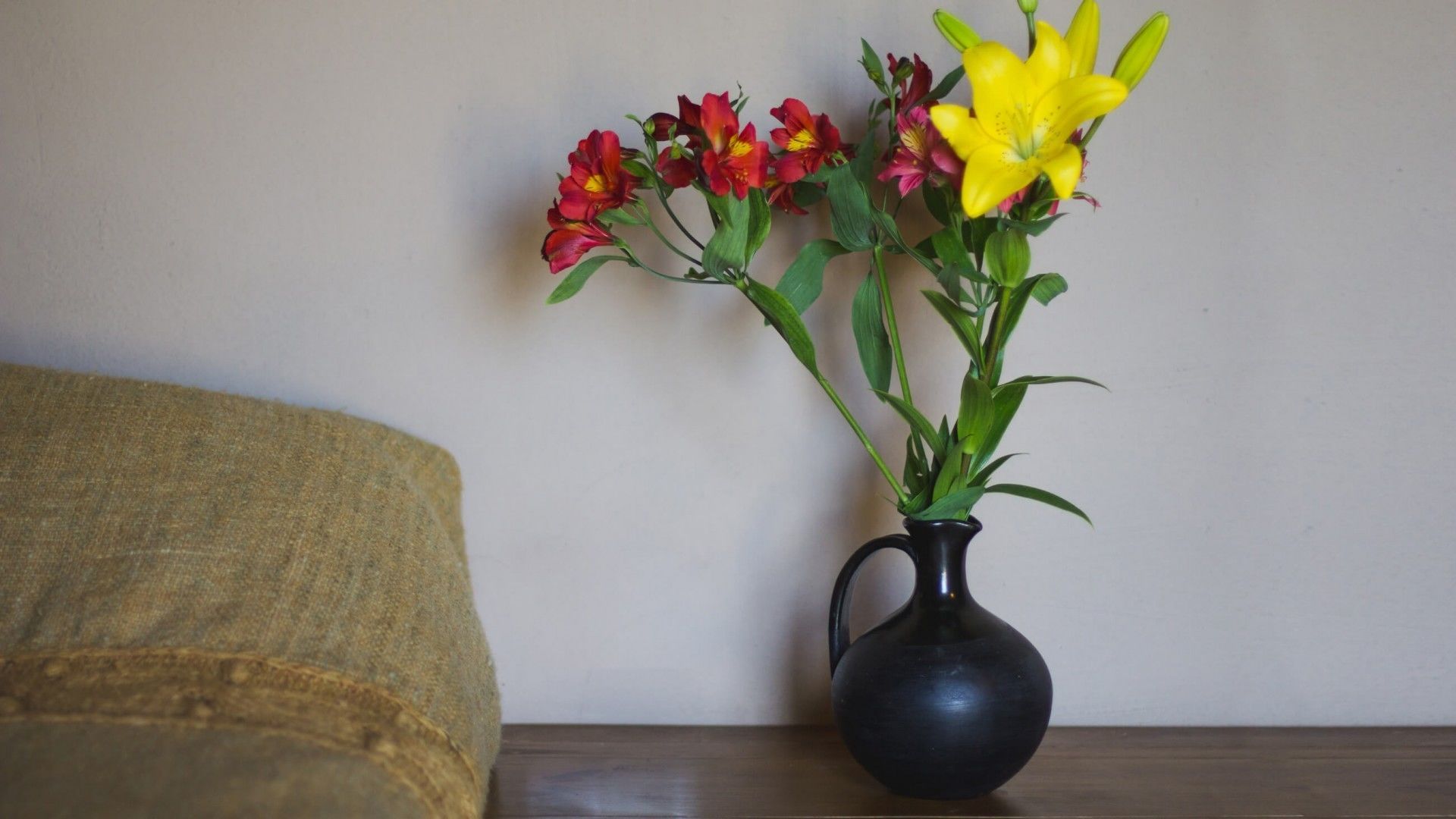 A black vase with flowers on a wooden table against a plain wall.