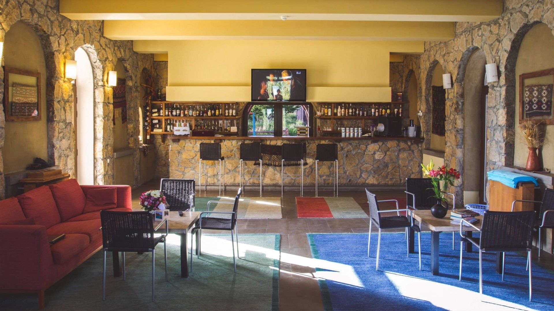 Eine Bar mit Steinwänden, einem Fernseher und einer Auswahl an Flaschen. Mehrere Stühle und ein rotes Sofa sind vorhanden.