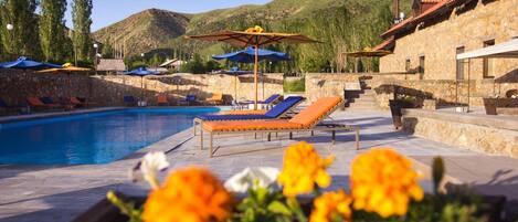 Zona de piscina con sillones de descanso y sombrillas, rodeada de muros de piedra y un paisaje montañoso.
