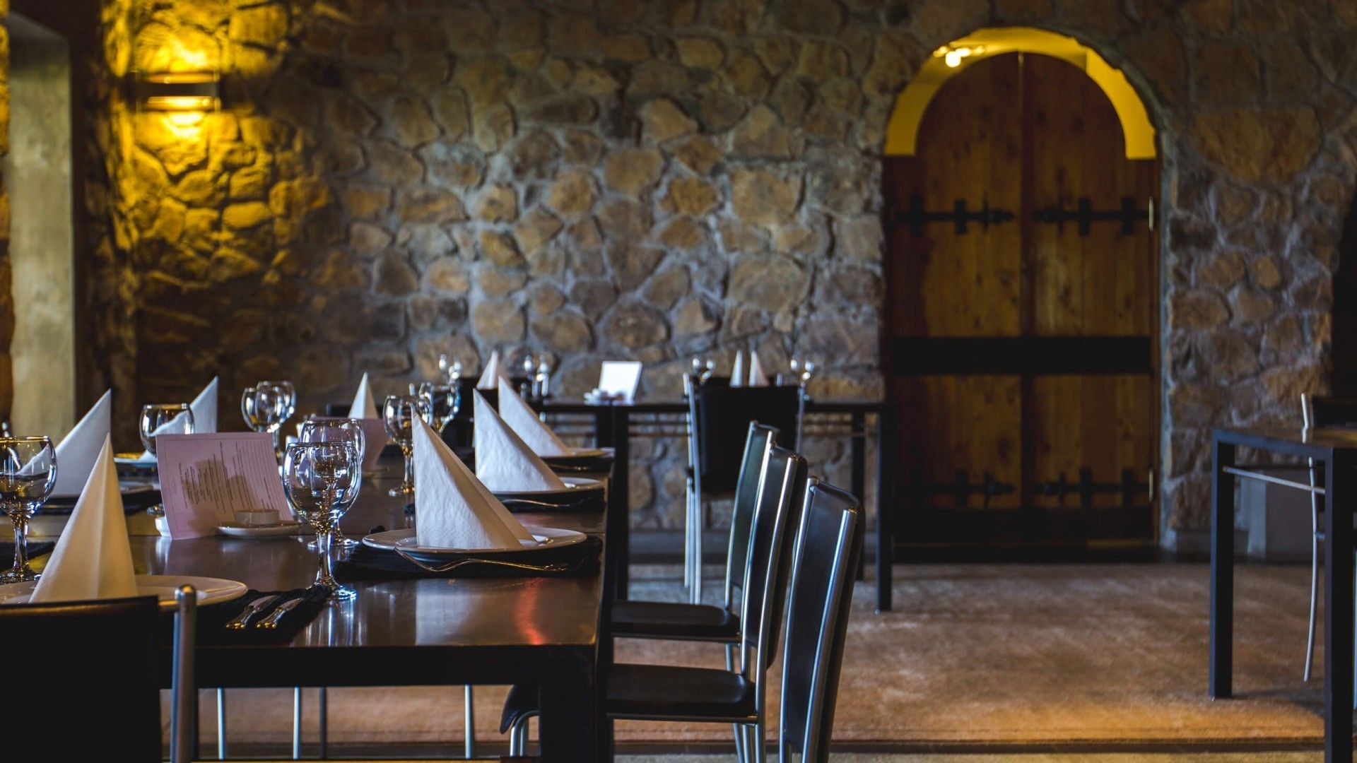 Ein Restaurant mit Steinwänden und einer Holztür. Tische sind mit Weingläsern und Speisekarten gedeckt.