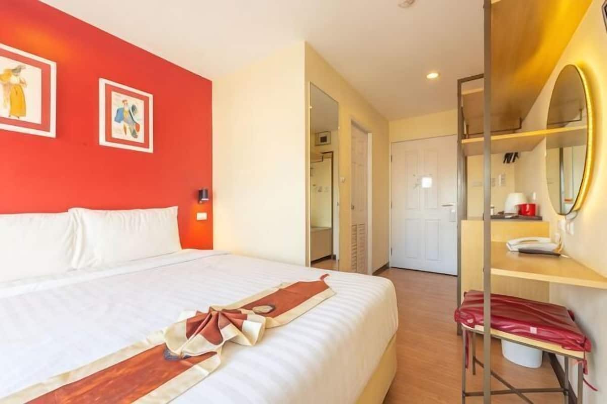A hotel room with a bed, a desk, a mirror, and a red wall.