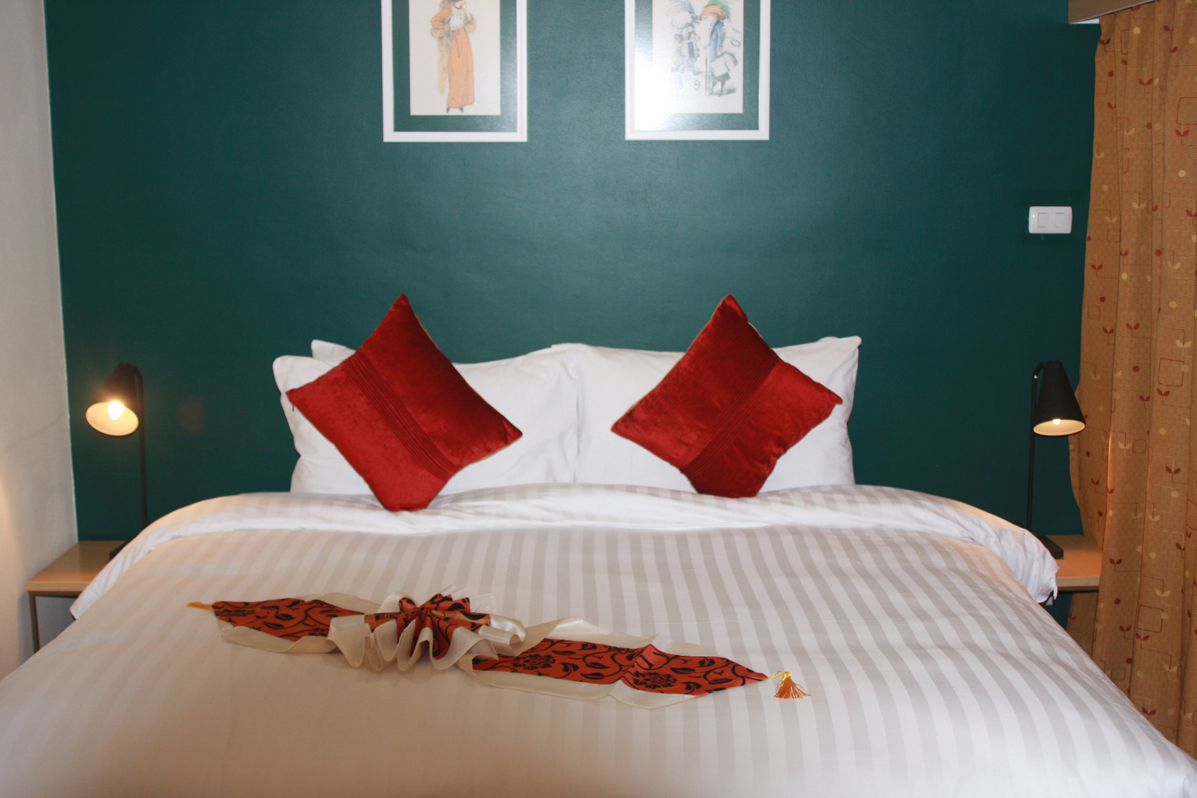 A neatly made bed with white bedding and red pillows, a decorative bow, and two framed artworks on the teal wall.