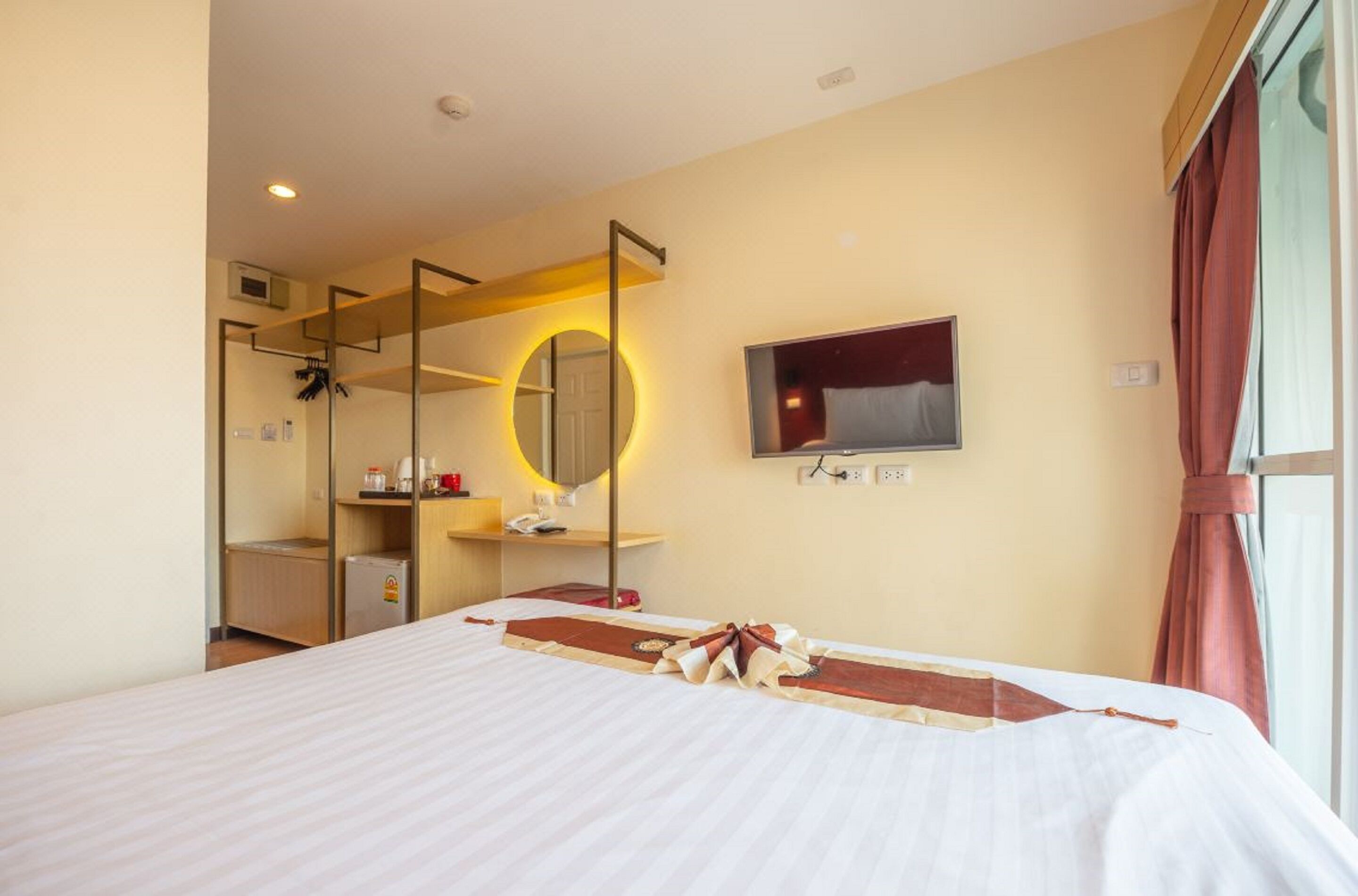 A hotel room with a bed, a TV, a mirror, a small kitchenette, and a window with curtains.