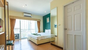 A hotel room with a bed, a desk, a chair, a window with curtains, an air conditioner, and a door with louvered panels. - Klean Residence Hotel (Bangkok)