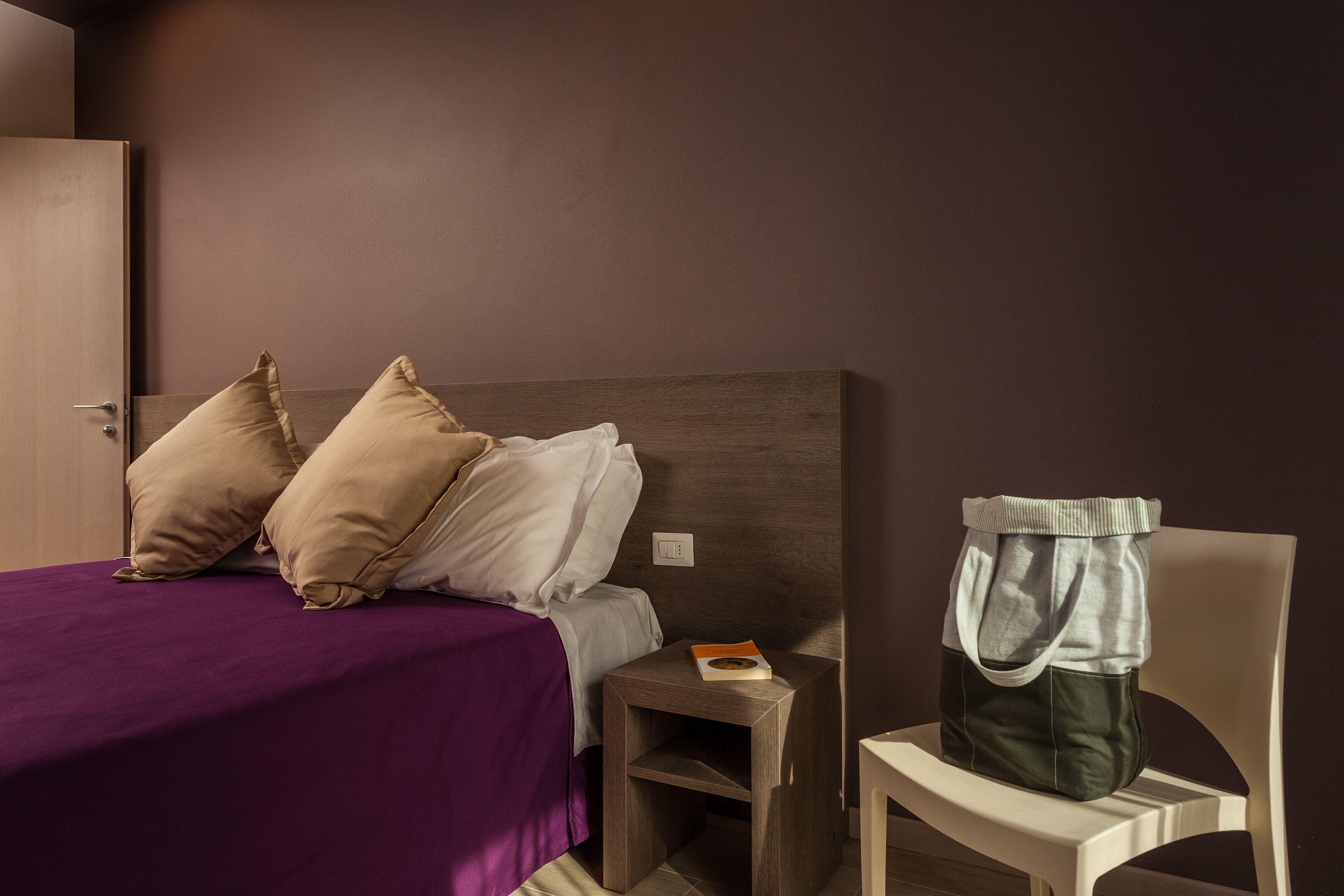 A bedroom with a bed, pillows, a nightstand, a chair, and a bag.