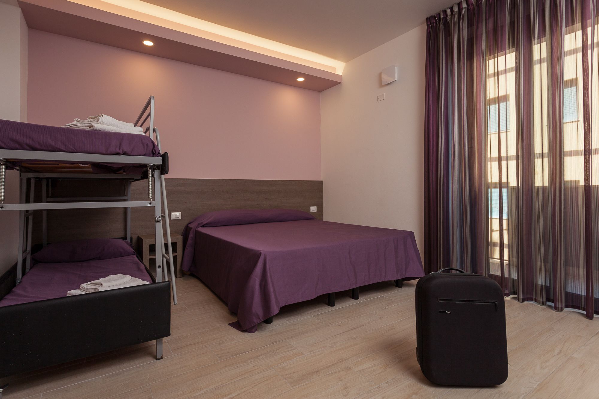 Une chambre d’hôtel avec des lits superposés, un lit simple, une valise et une fenêtre avec des rideaux.