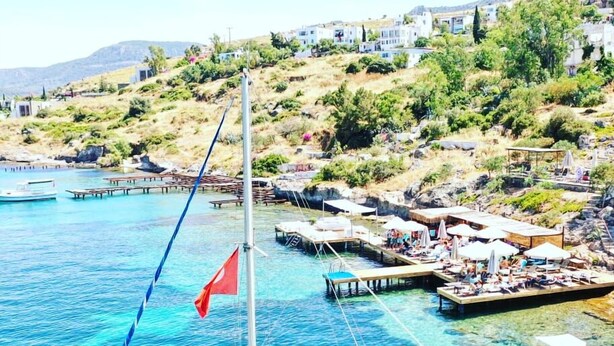 A sailboat docked at a marina with clear blue water, surrounded by a small coastal village with houses and greenery. - Beste Hotel (Bodrum)