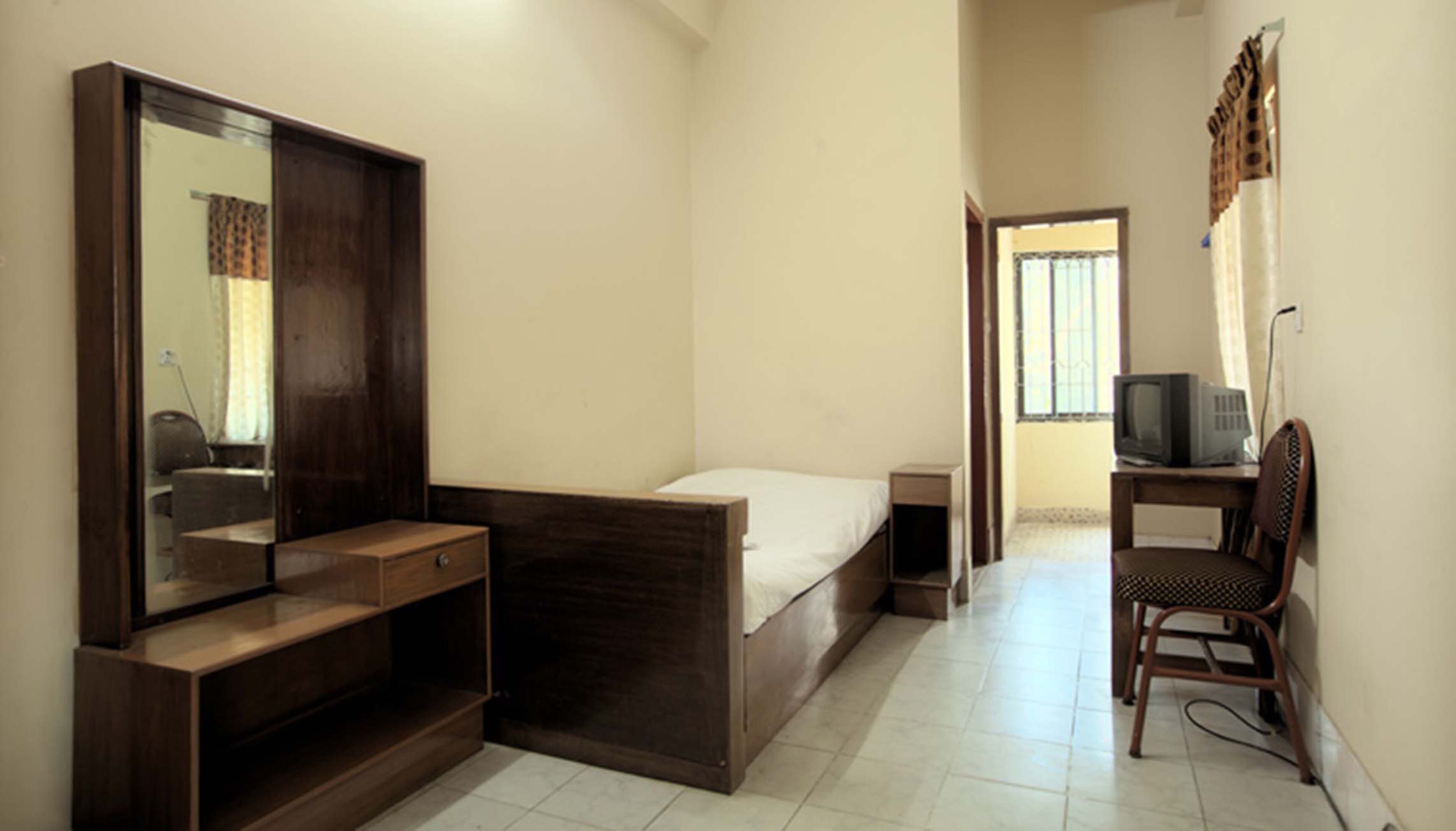 A bedroom with a bed, a wooden wardrobe, a desk with a chair, a television, and a window.