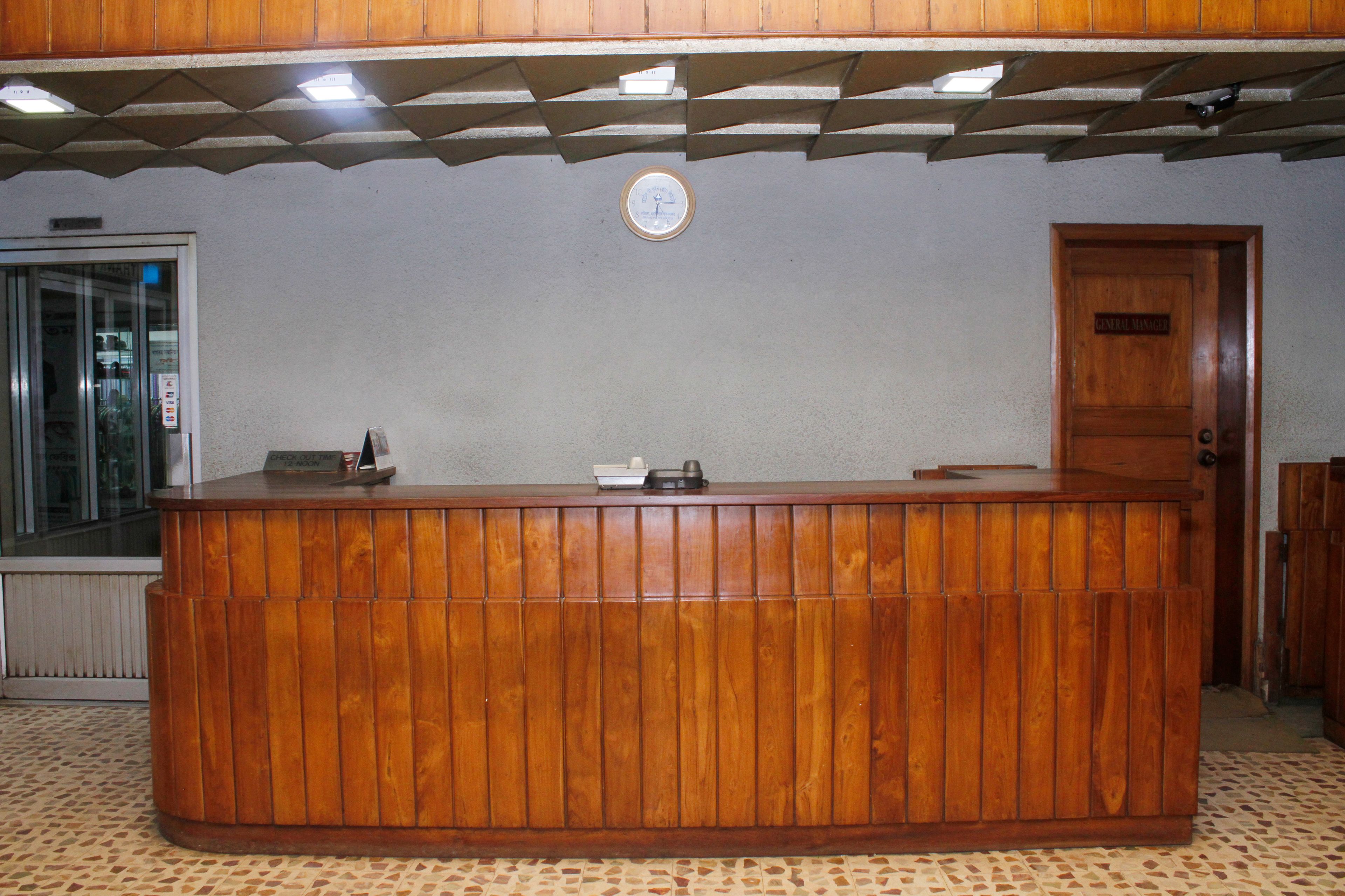 Egy fából készült recepciós pult, a falon egy óra, egy „Recepció” felirattal ellátott ajtó és egy zsaluzatos ablak.