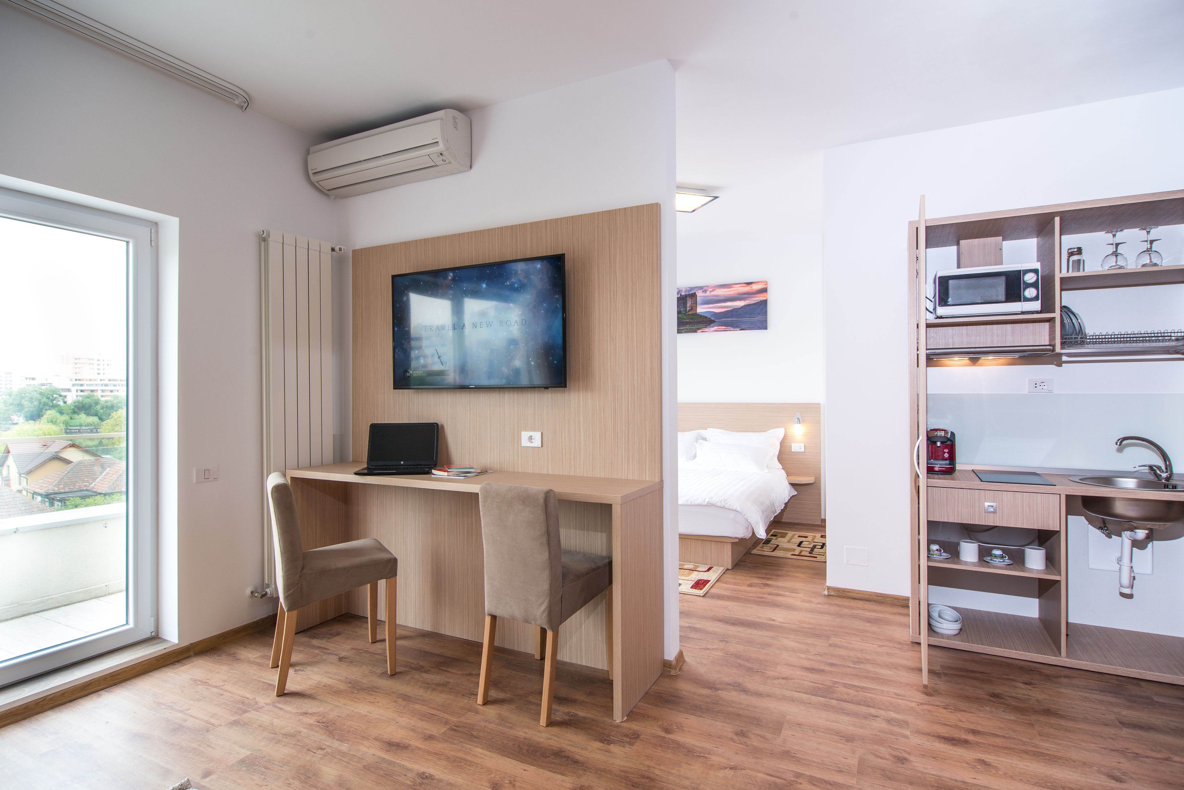 Una habitación de hotel moderna con un escritorio de madera, un televisor de pantalla plana, una cama con ropa de cama blanca, una cocina integrada con microondas y un amplio ventanal con vistas a la ciudad.