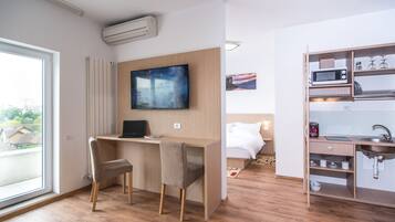 Una habitación de hotel moderna con un escritorio de madera, un televisor de pantalla plana, una cama con ropa de cama blanca, una cocina integrada con microondas y un amplio ventanal con vistas a la ciudad.