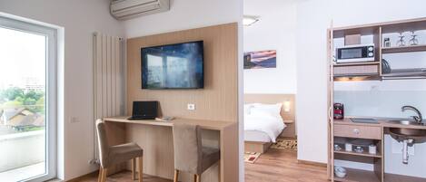 A modern hotel room with a wooden desk, a flat-screen TV, a bed with white bedding, a built-in kitchen with a microwave, and a large window offering a city view.