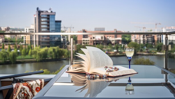 A book and a glass of wine on a glass table with a view of a river and buildings in the background.