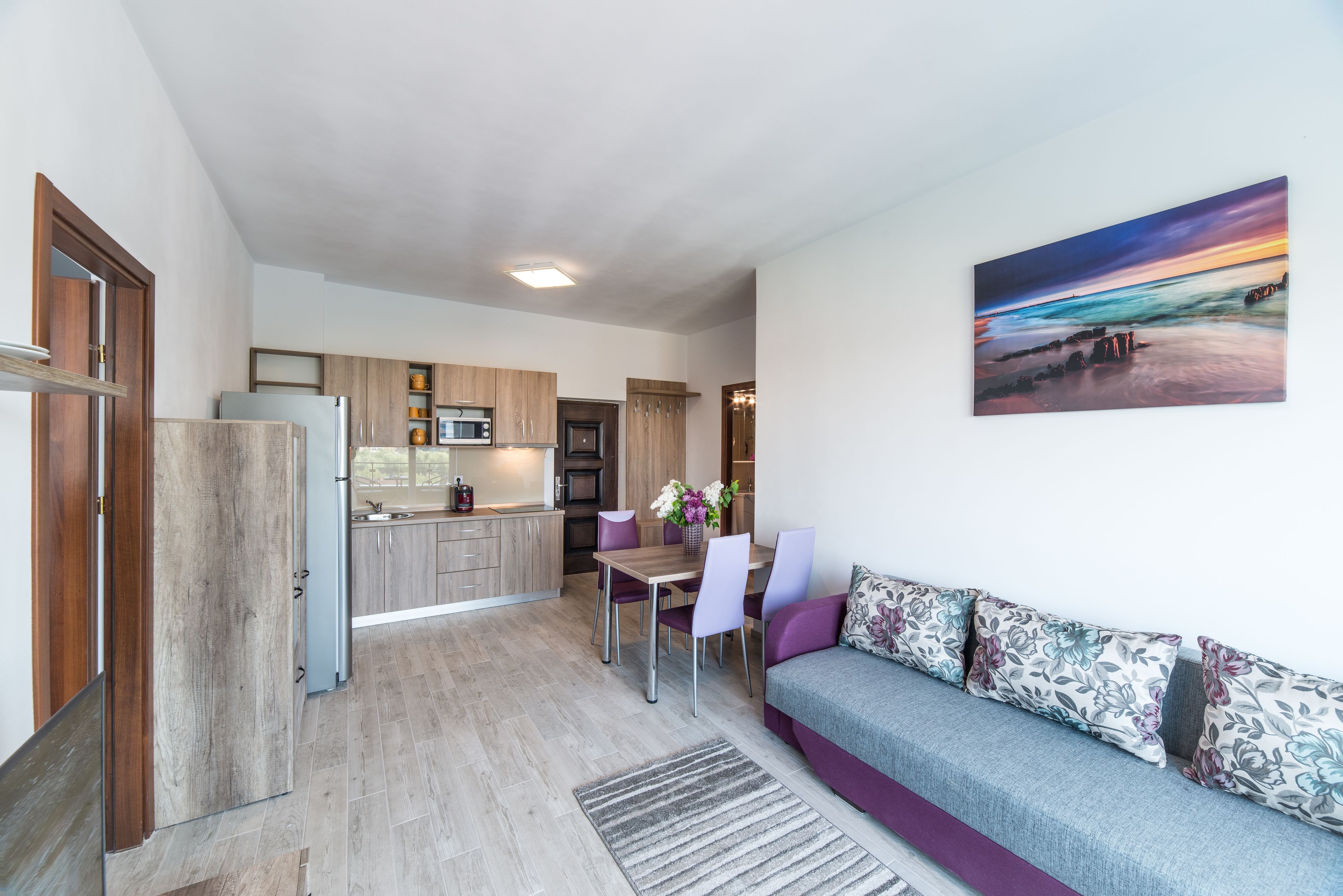 A modern kitchen with wooden cabinets, a dining area with purple chairs, and a gray sofa with floral cushions.