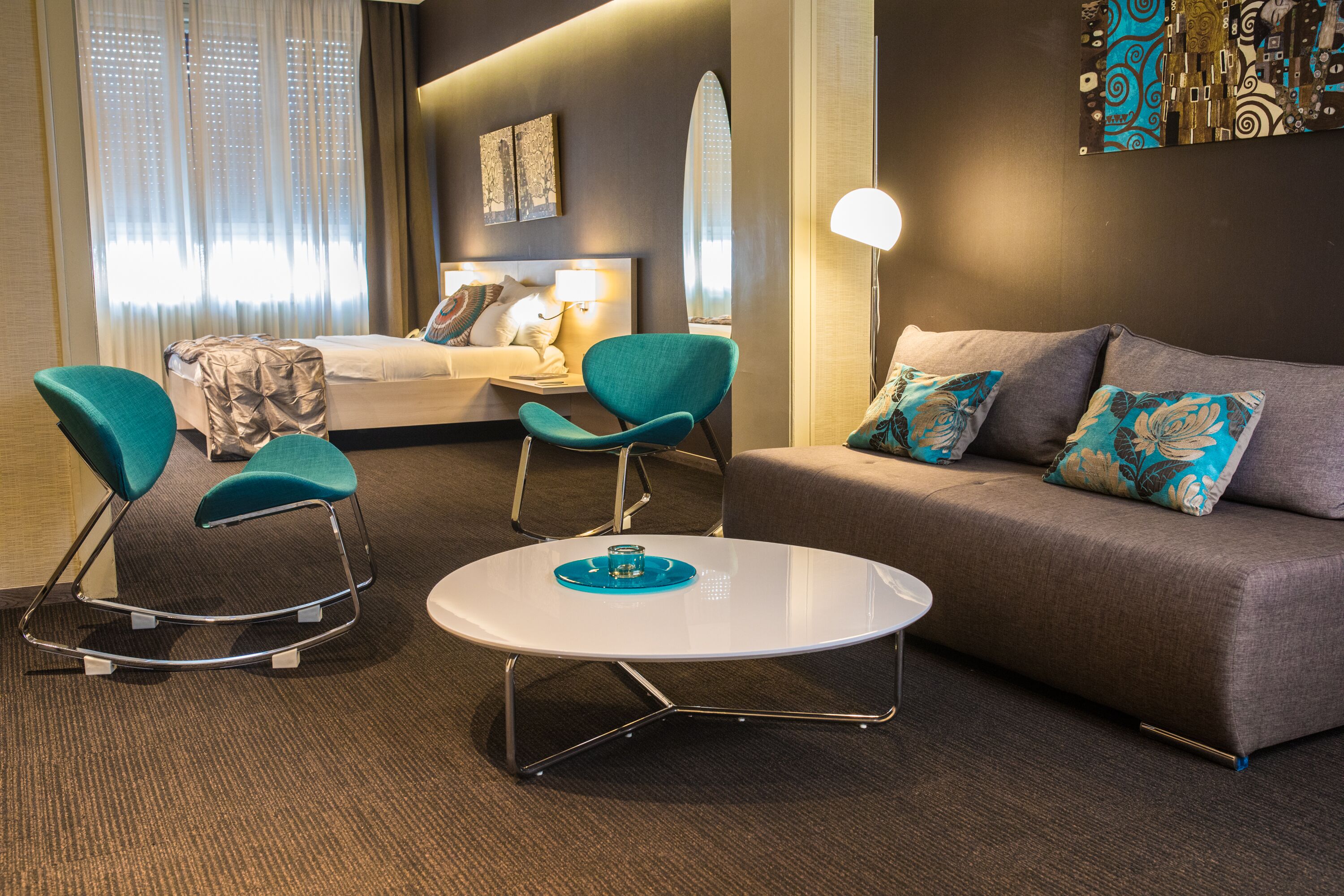 A hotel room with a bed, two teal chairs, a white coffee table, a grey sofa, and a large abstract painting on the wall.