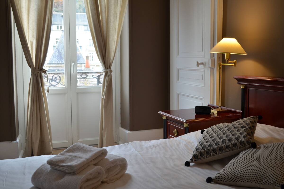 A hotel room with a bed, a bedside lamp, a nightstand, and a view of the outside through a window with curtains.
