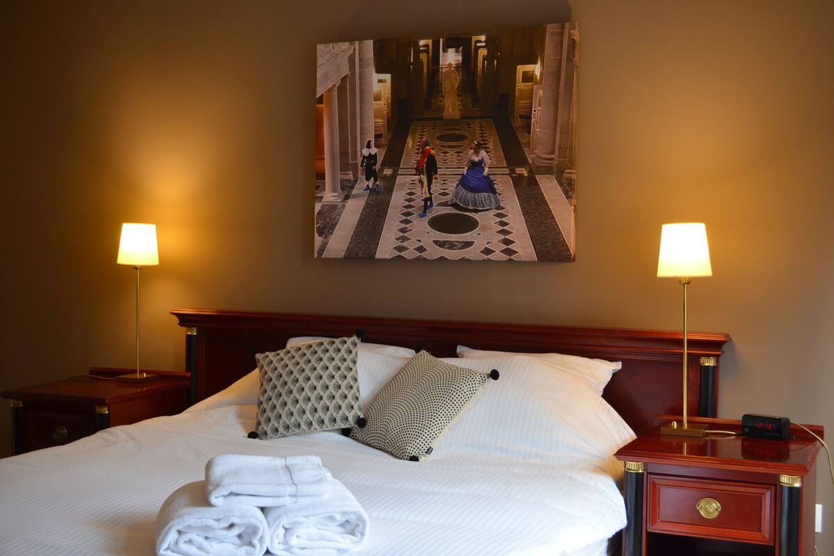 A hotel room with a large bed, two bedside tables, a nightstand, and a painting of a hallway.