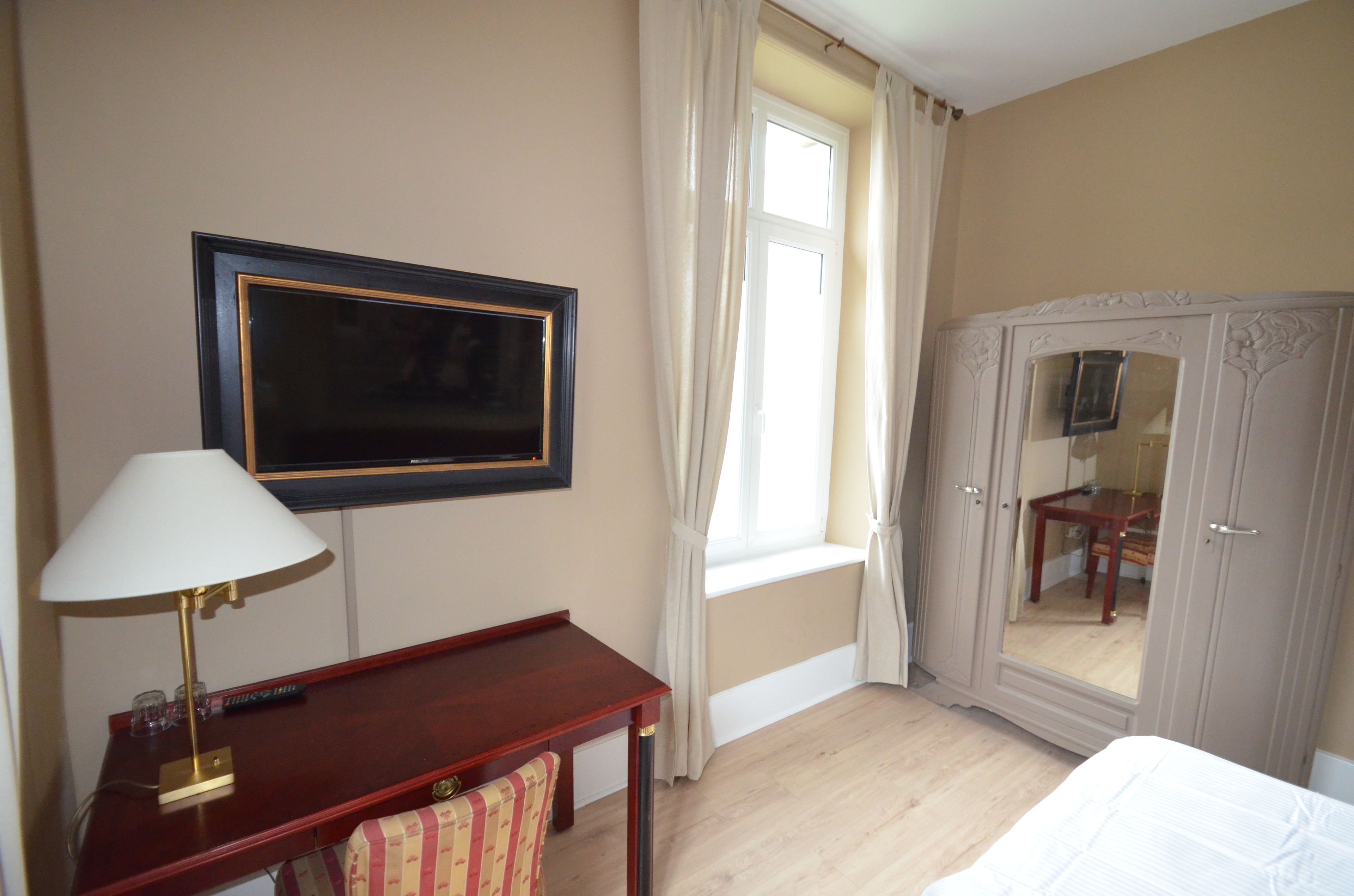 Une chambre d’hôtel avec un lit, un bureau avec une lampe, un téléviseur fixé au mur, une armoire et une fenêtre avec des rideaux.