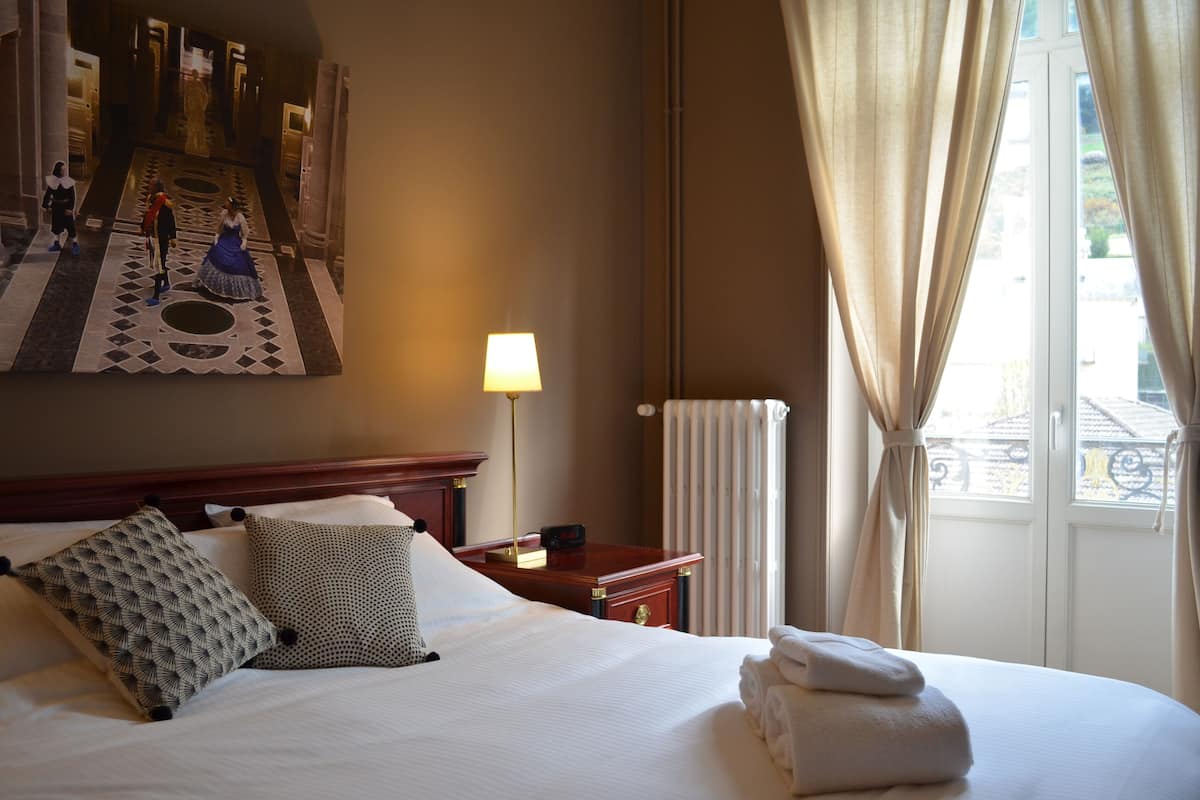 A hotel room with a large bed, a nightstand with a lamp, a painting on the wall, and a window with curtains.