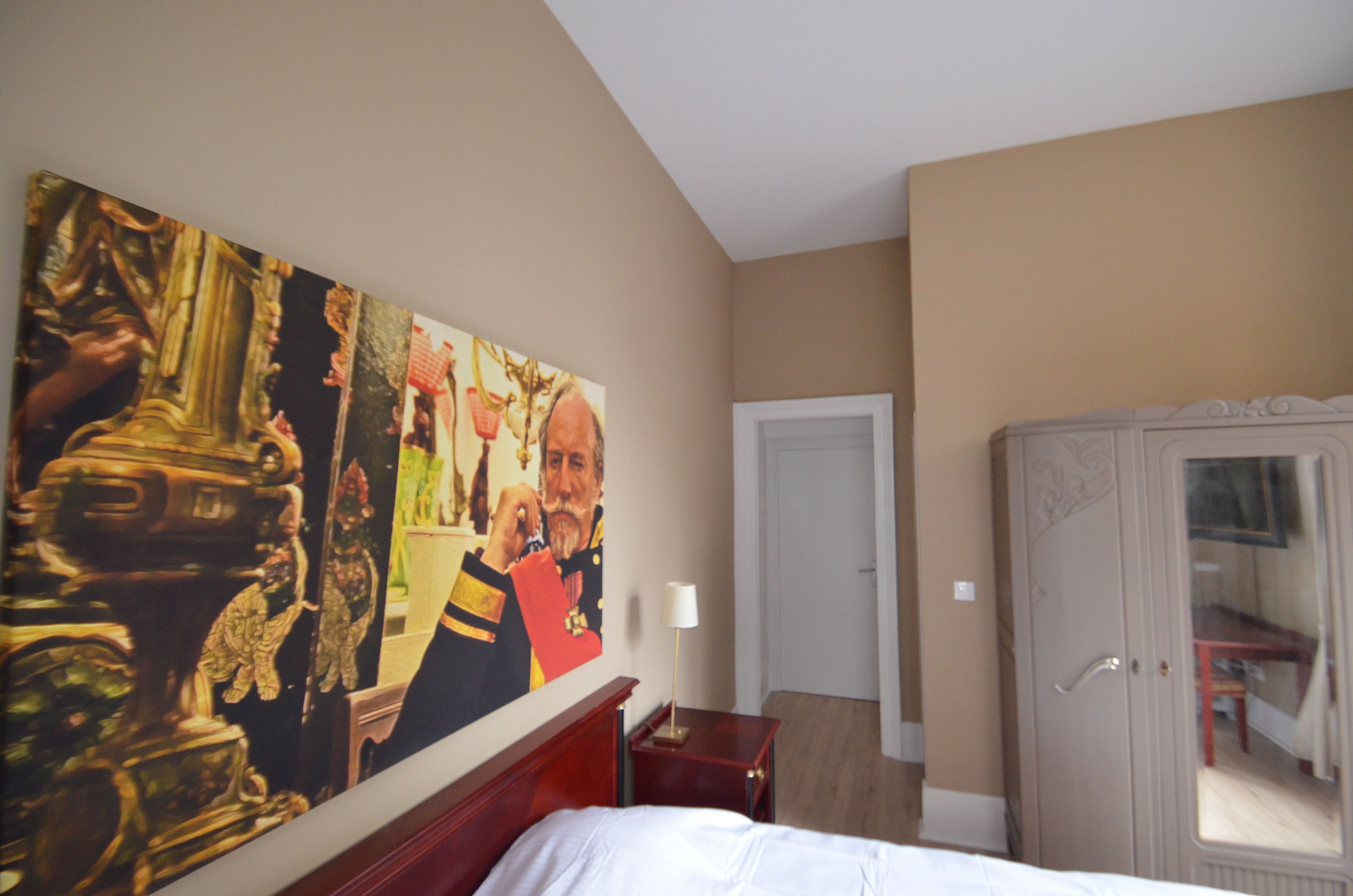 Ein Hotelzimmer mit großem Gemälde an der Wand, einem Bett, einem Kleiderschrank und einem Schreibtisch mit Lampe.