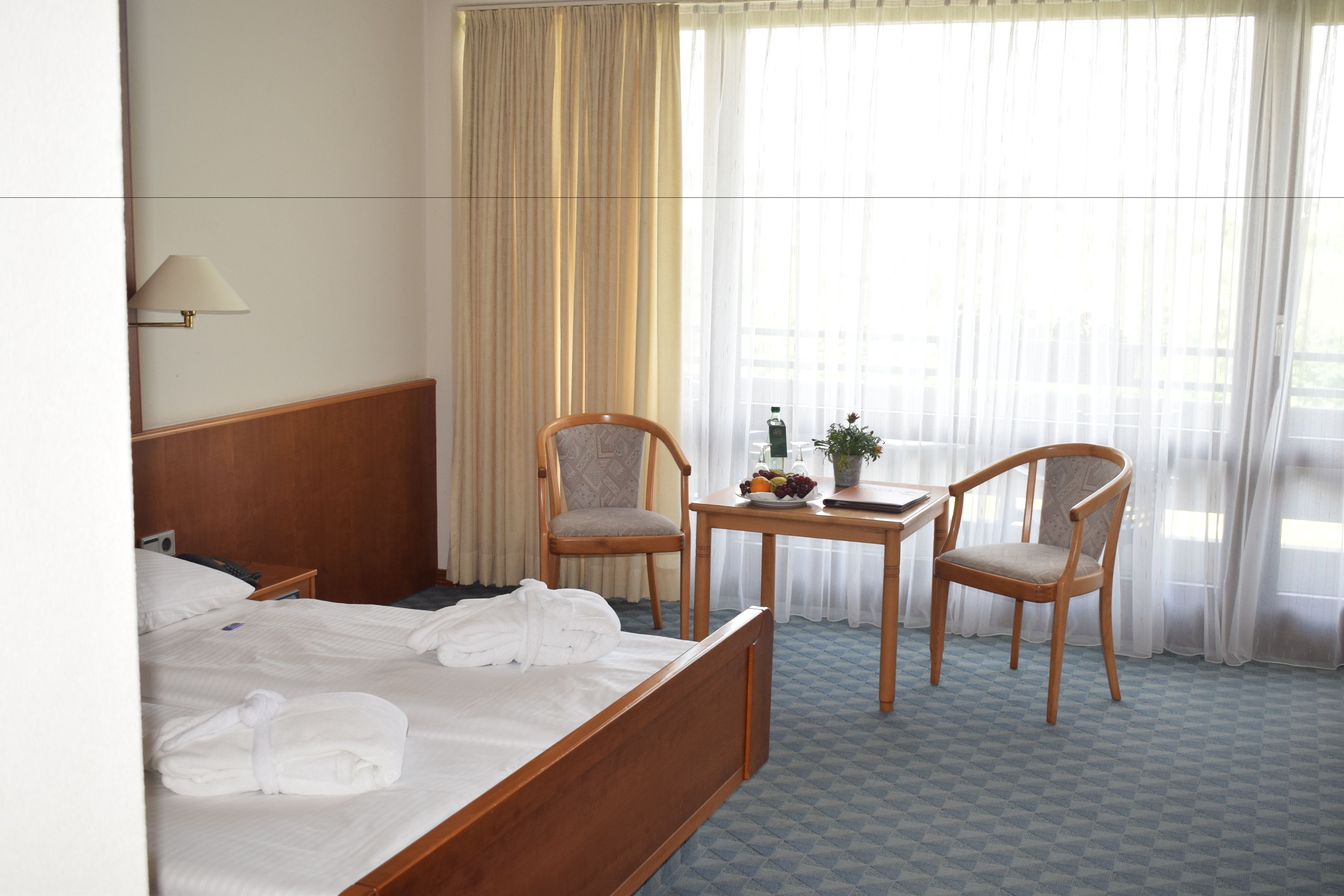 A hotel room with a bed, two chairs, a small table with a fruit bowl and a bottle, and a window with sheer curtains.
