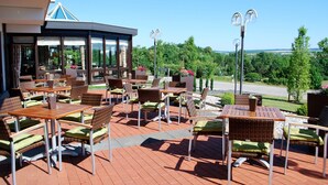 An outdoor seating area with wicker chairs and tables, a covered section with large windows, and a view of greenery and a road in the background.