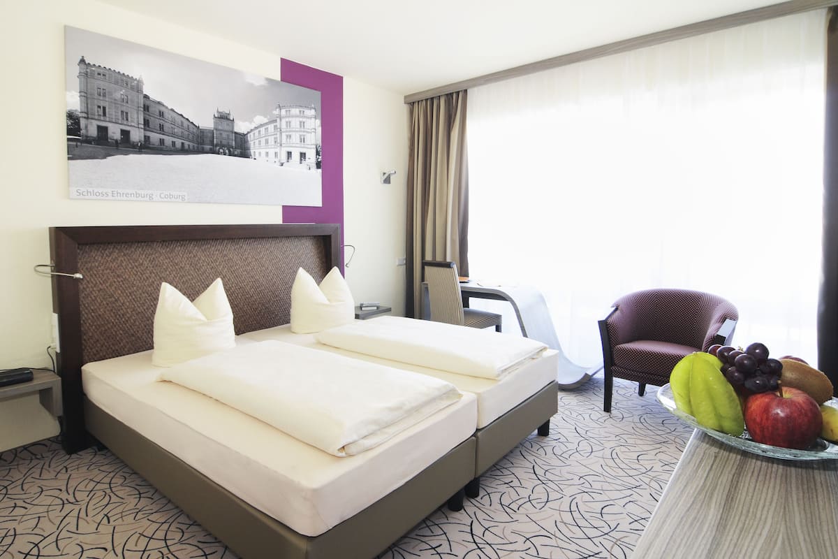A hotel room with two beds, a chair, a table with fruit, and a framed picture on the wall.
