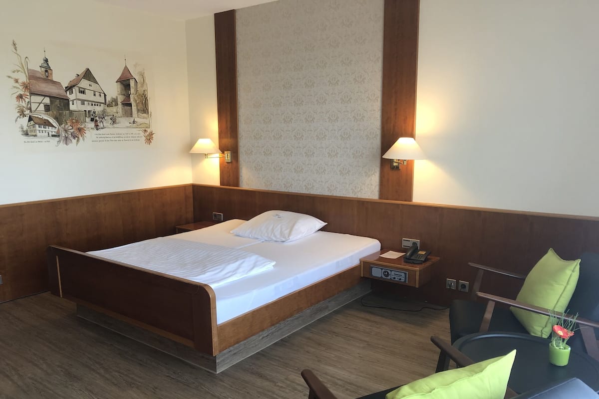 A hotel room with a wooden bed, two bedside lamps, a nightstand with a phone, and a chair with a green cushion.