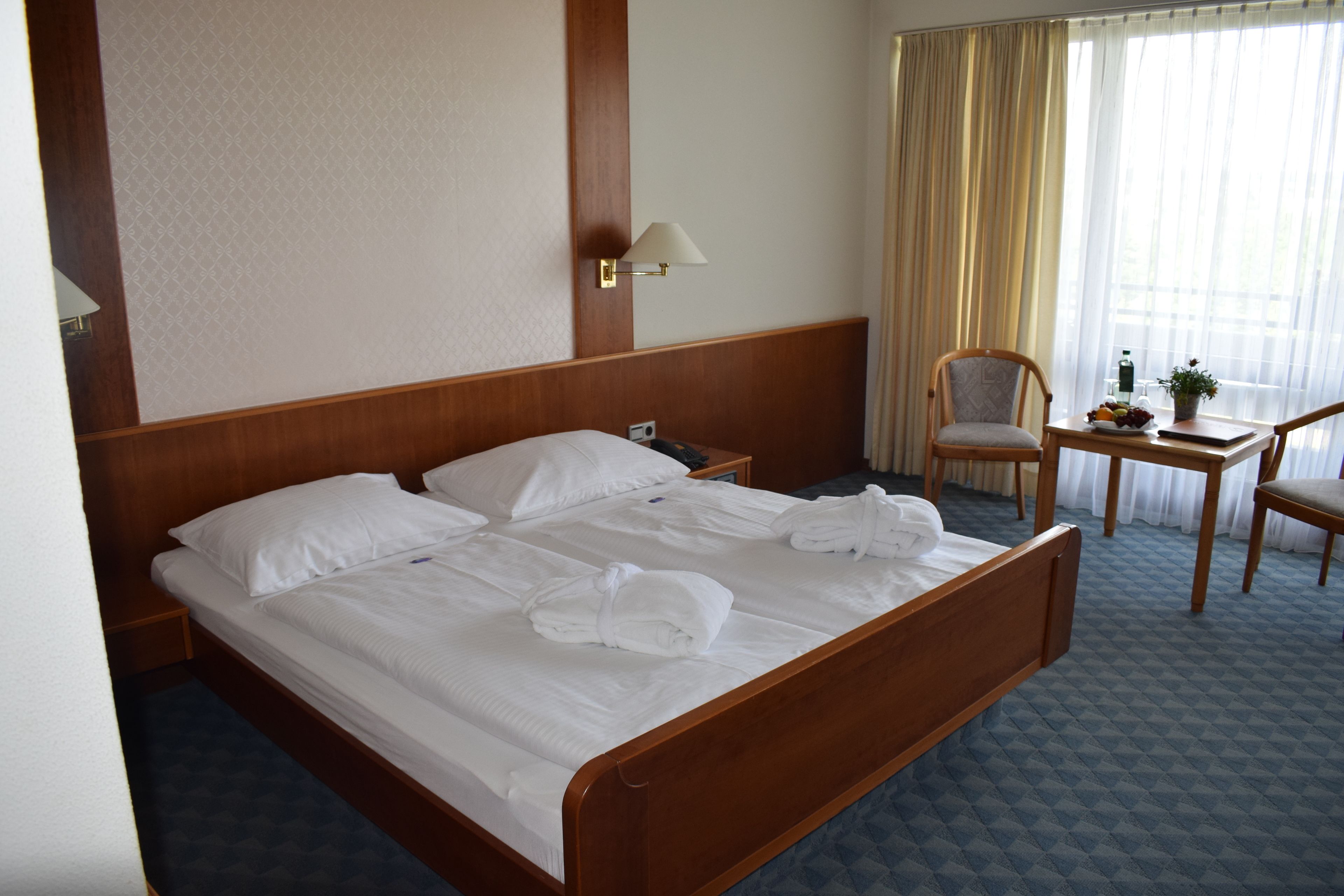 Ein Hotelzimmer mit einem hölzernen Bett, einem Nachttisch, einem Sessel, einem kleinen Tisch mit Pflanze und einem Fenster mit Vorhängen.